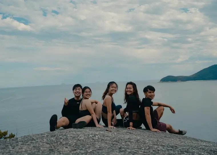 Five young adults sitting shoulder to shoulder on a rock above the sea, smiling at the camera