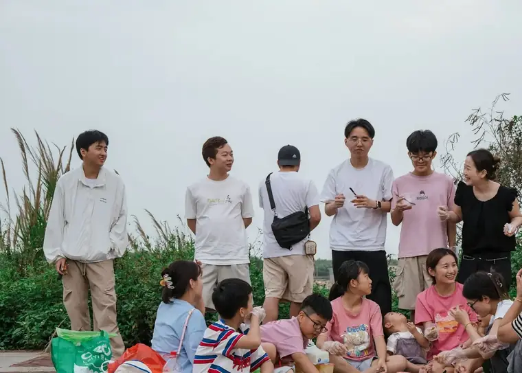Adults standing and children sitting together outdoors beside tall grass, sharing food
