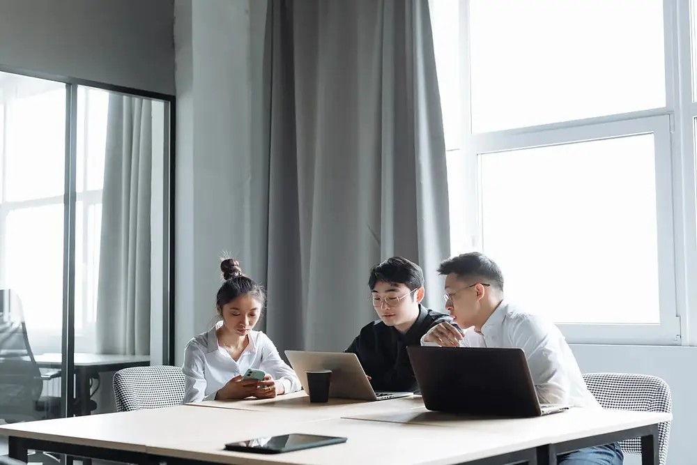 Three colleagues conferring over a laptop at a table in a bright office