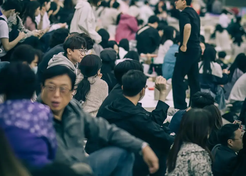 A dense indoor crowd of all ages seated close together, some talking, some eating